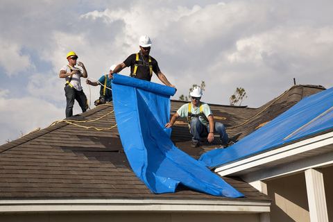 Roofers in safety harnesses unrolling blue sheeting over a storm-damaged shingle roof