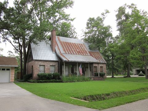 Brick cottage with a rust-streaked metal roof and covered porch beneath tall shade trees