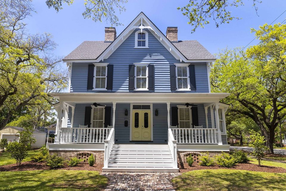 Blue two-story wooden house with a gray shingle roof, wide front porch and live oaks