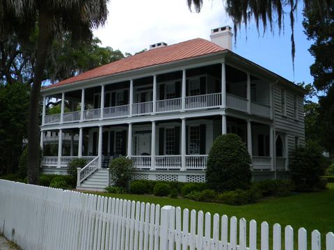 Two-story white house with a red metal roof and double porches behind a picket fence