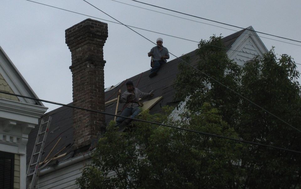 Two roofers working on a steep dark roof beside a tall brick chimney